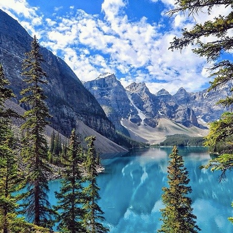Turquoise mountain lake with pine trees and snow-capped peaks under a partly cloudy sky