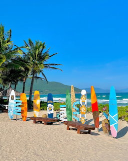Colorful surfboards lined up on a sandy beach with palm trees, ocean waves, and mountains in the background.