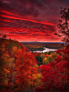 Brilliant red autumn trees frame a lake view under a fiery sunset sky with dramatic red clouds