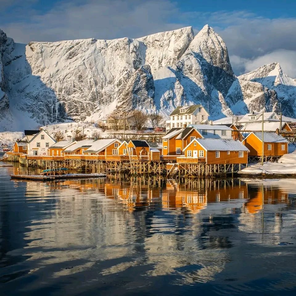 Snowy Norwegian fishing village with colorful houses, calm water reflections, and steep mountains