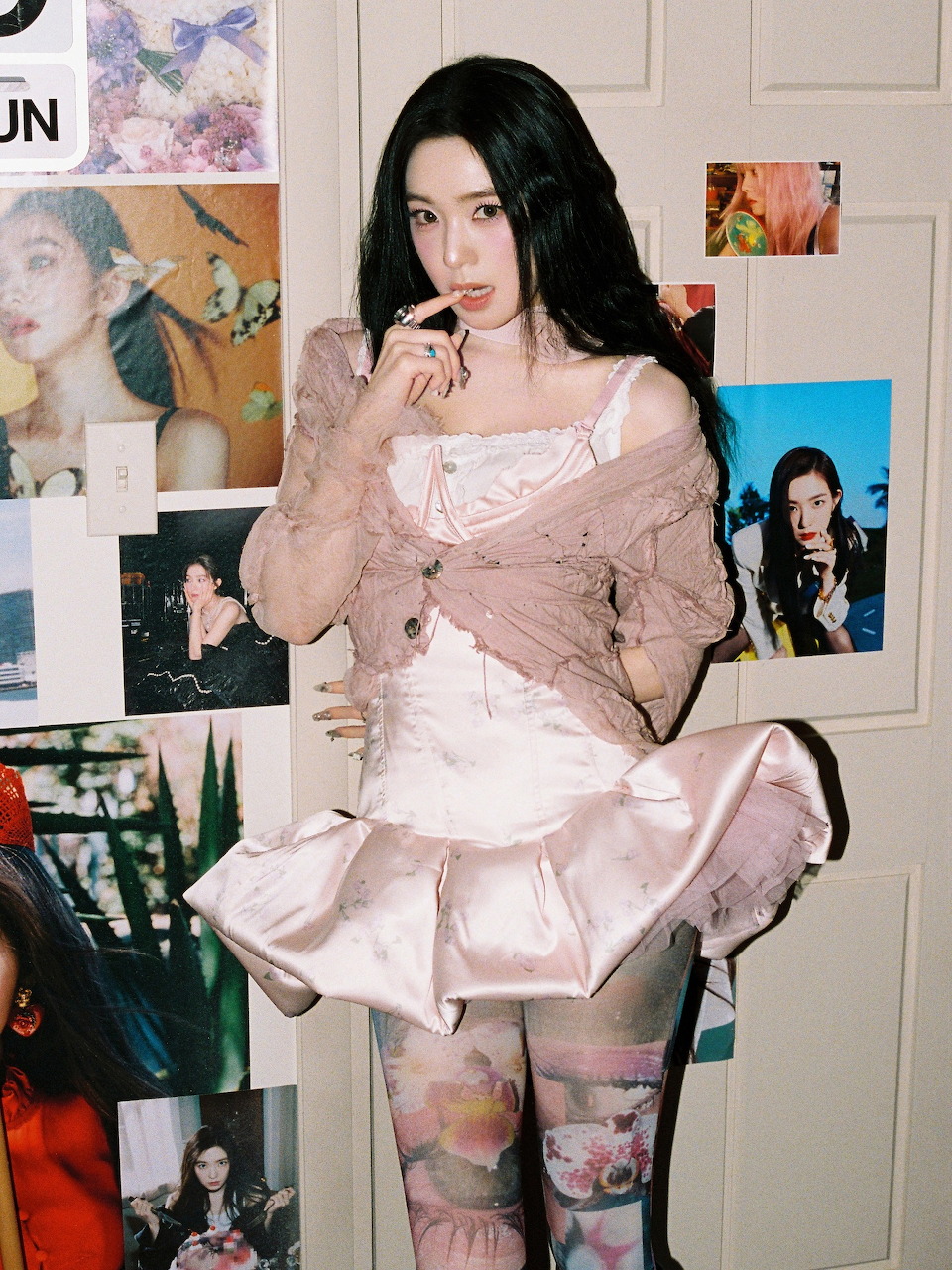 Woman with long black hair in pink ruffled top and skirt posing in front of a photo-covered wall
