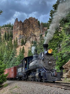 Black steam train with red railcars traveling through a forested rocky canyon