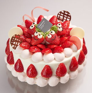 Round cake topped with strawberries, white cream, chocolate pieces, and strawberry-shaped characters with googly eyes