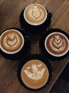 Overhead view of four black cups of latte with leaf latte art on a wooden table