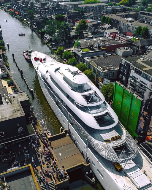 Aerial view of a large white luxury yacht passing through a narrow Amsterdam canal lined with buildings and crowds