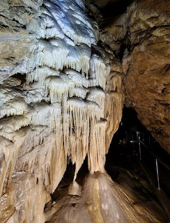 White and tan stalactites hanging inside a rocky cave with a dark passage and railing