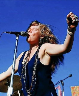 Janis Joplin singing on stage into a microphone with her arm raised against a blue sky