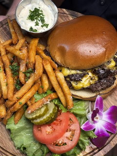Cheeseburger and seasoned fries on a wooden plate with pickles, tomato, lettuce, sauce, and orchid garnish.