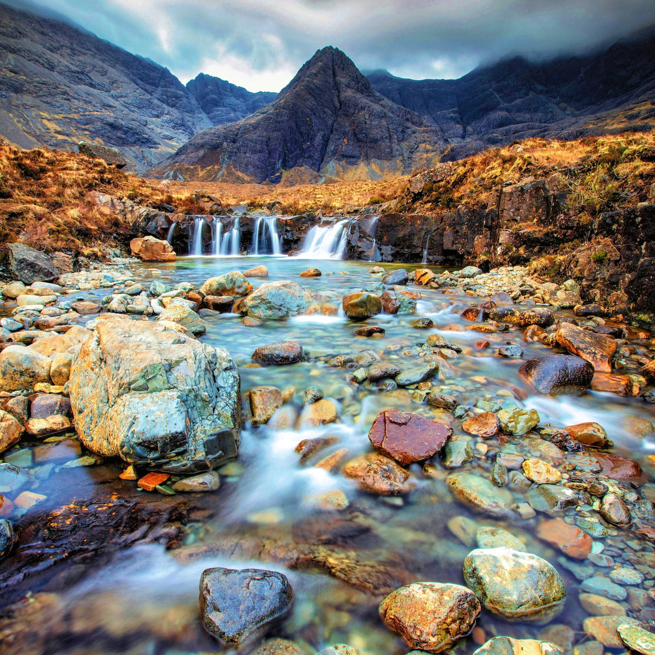 Waterfall cascading over colorful rocks with dramatic mountain peaks and moody clouds in Scottish Highlands