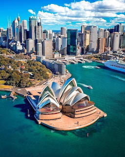 Aerial view of Sydney Opera House, harbour, and city skyline with blue water and clouds