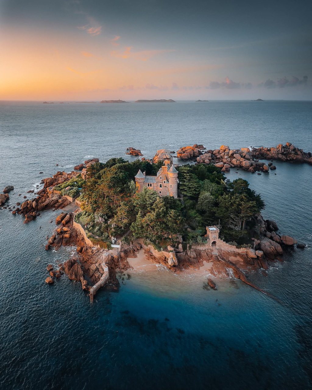 Aerial view of lighthouse on small rocky island surrounded by turquoise ocean at sunset