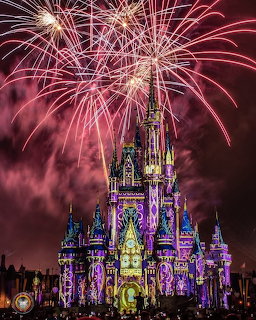 Fireworks bursting above an illuminated fairy tale castle at night with pink and purple lighting