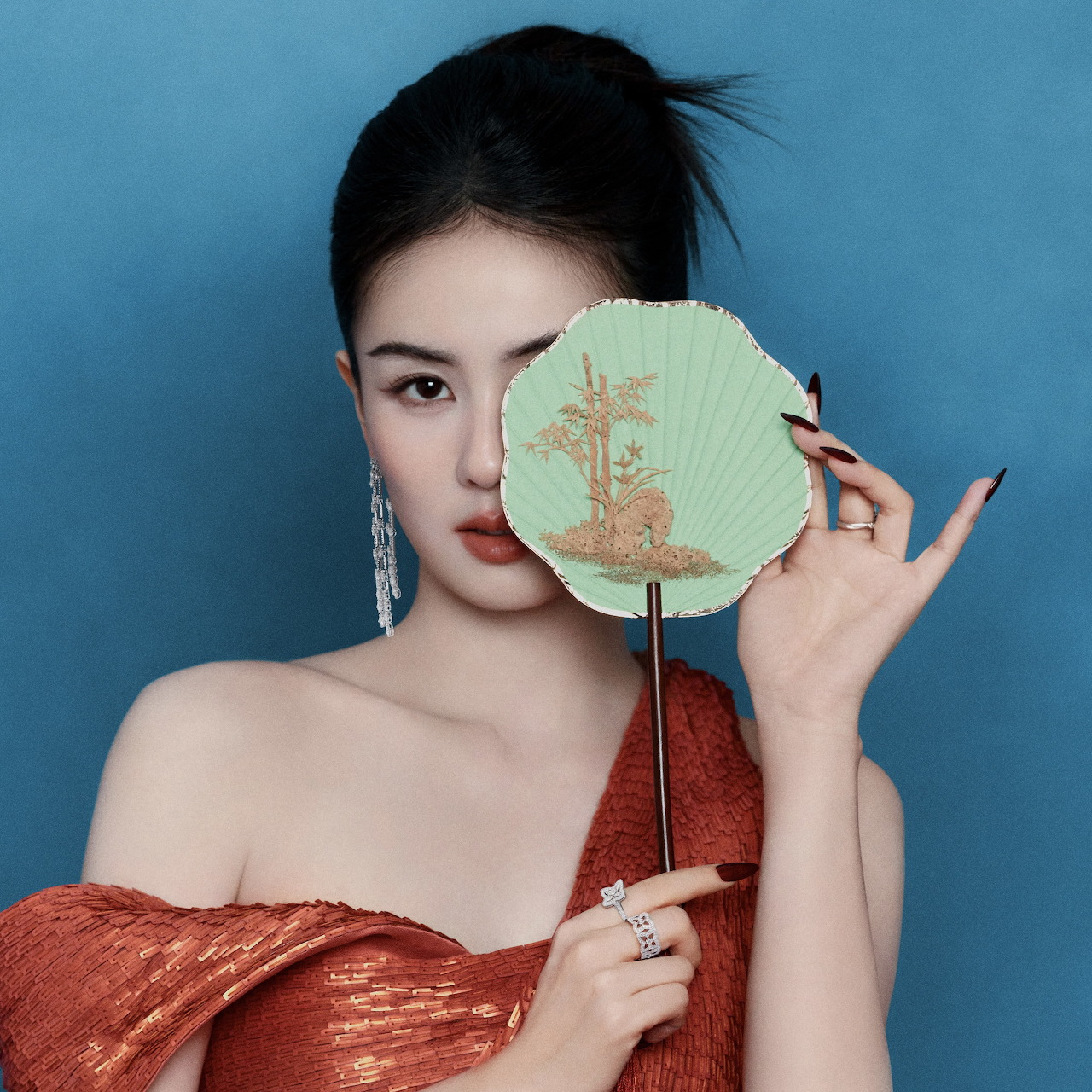 Woman in a red off-shoulder dress holding a round decorative fan in front of one eye