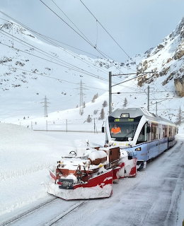 Modern train with red snowplow moving through deep snow in an alpine mountain setting