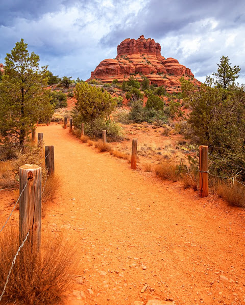 Desert trail with red sand leading to layered red rock butte, wooden fence posts and green shrubs along path, cloudy blue sky