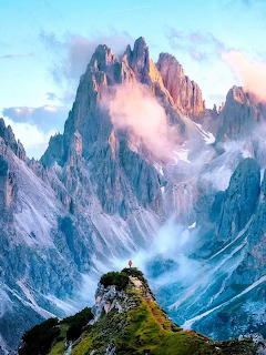 Jagged Dolomite mountain peaks towering above clouds with a lone hiker on a grassy ridge at sunset