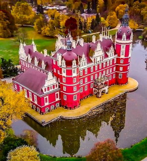 Red castle with spires and turrets surrounded by water and colorful autumn foliage