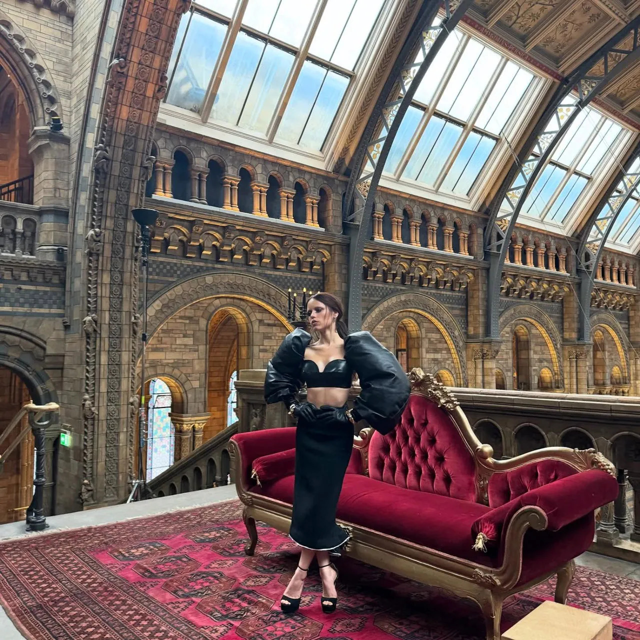 Woman in elegant black dress seated on burgundy Victorian sofa in ornate museum interior with arched skylights
