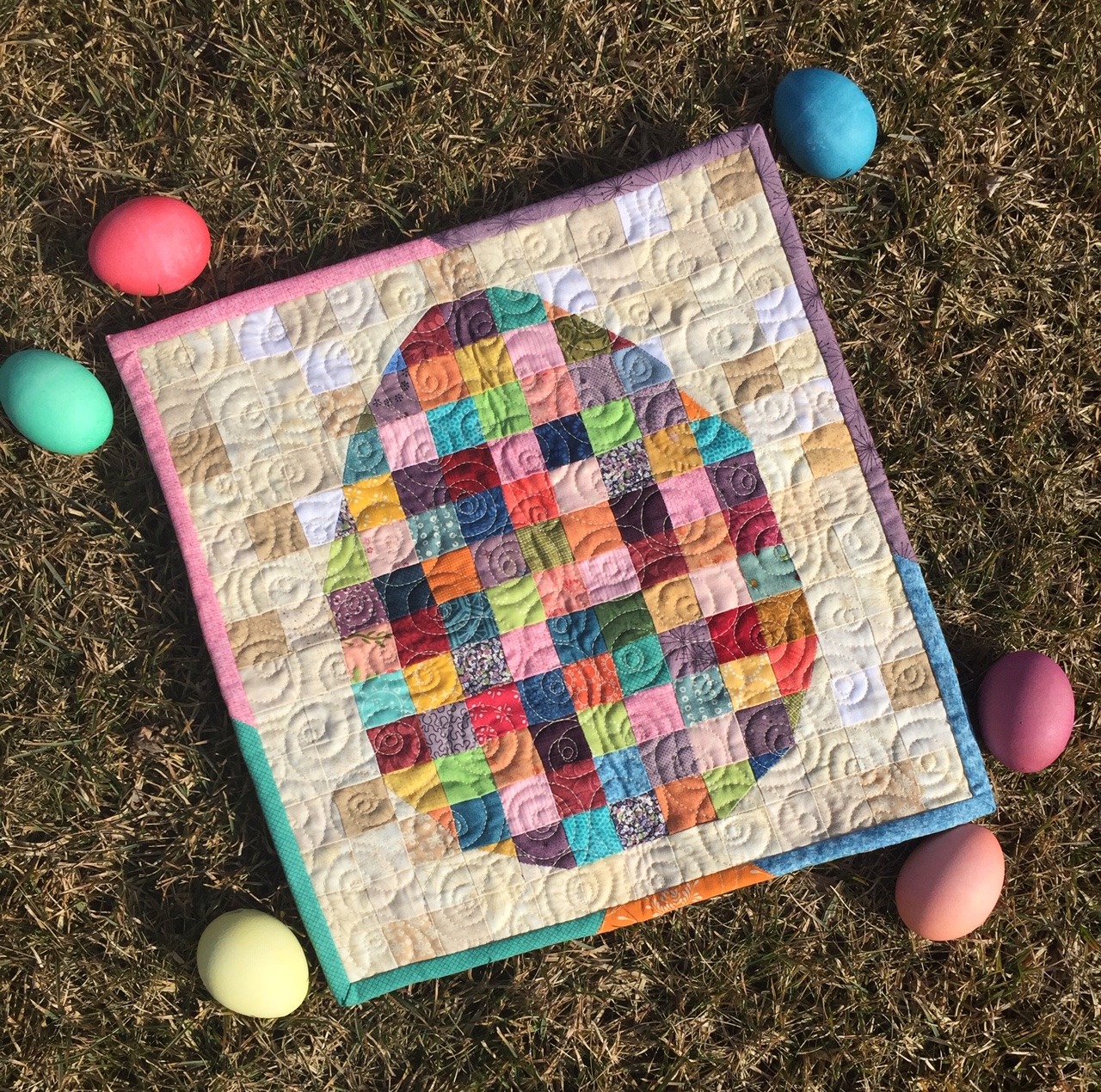 Handmade quilted wall hanging with pastel patchwork egg design surrounded by plastic Easter eggs on textured ground