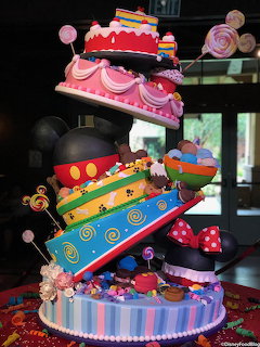 Tall colorful Mickey Mouse themed tiered cake with tilted layers, lollipops, and candy decorations