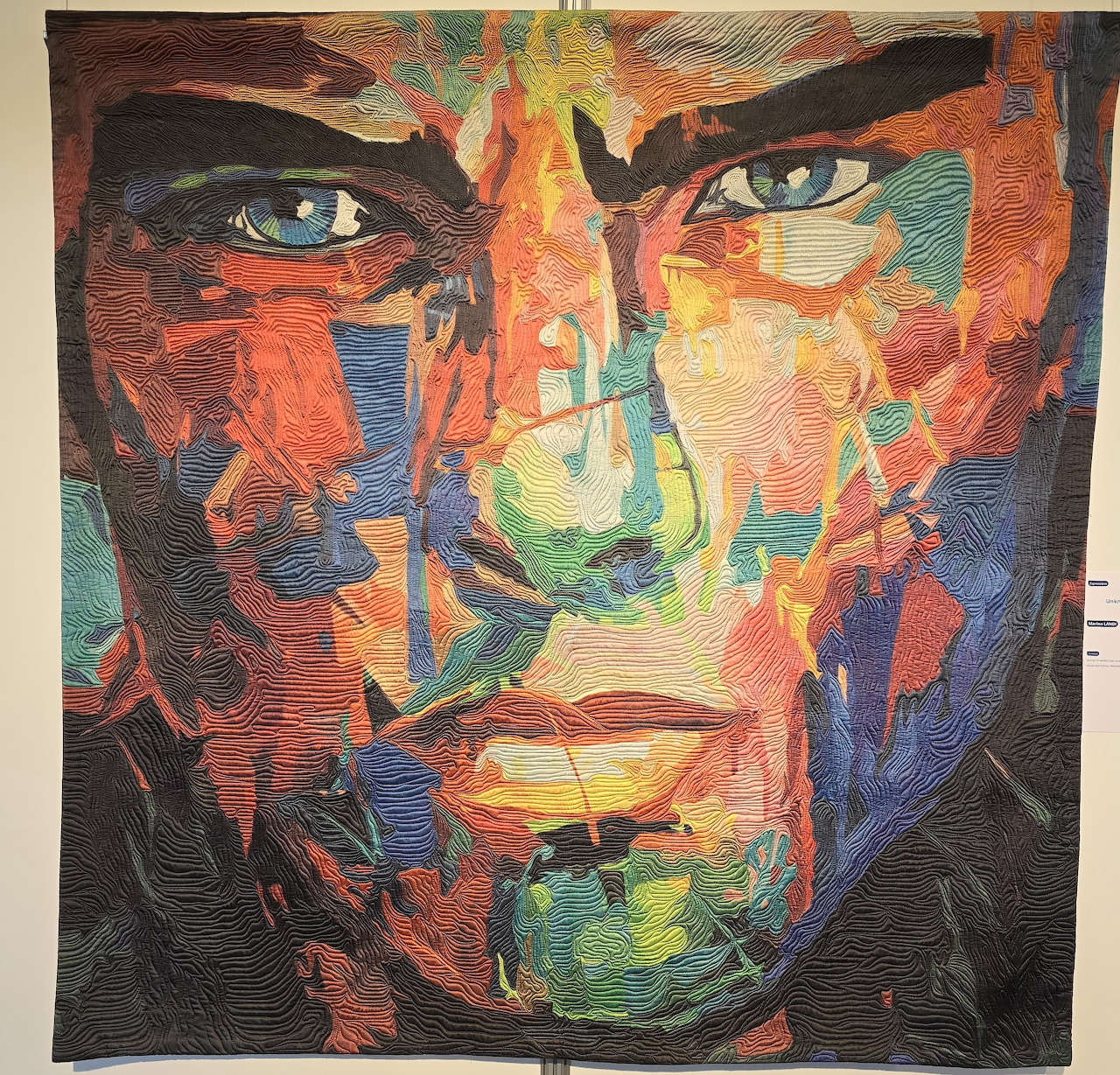 Colorful quilted portrait of a face made from patchwork fabric pieces in warm oranges, reds, blues, and greens