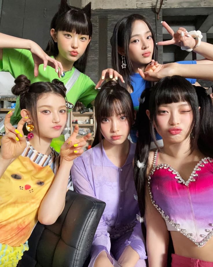 Five young women in colorful outfits posing together making playful hand gestures indoors