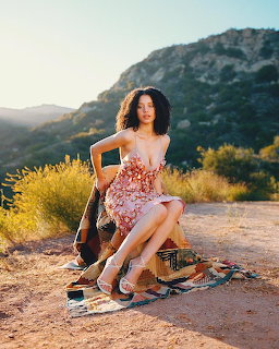 Woman in floral dress sitting on picnic blanket at mountain overlook during sunset with guitar