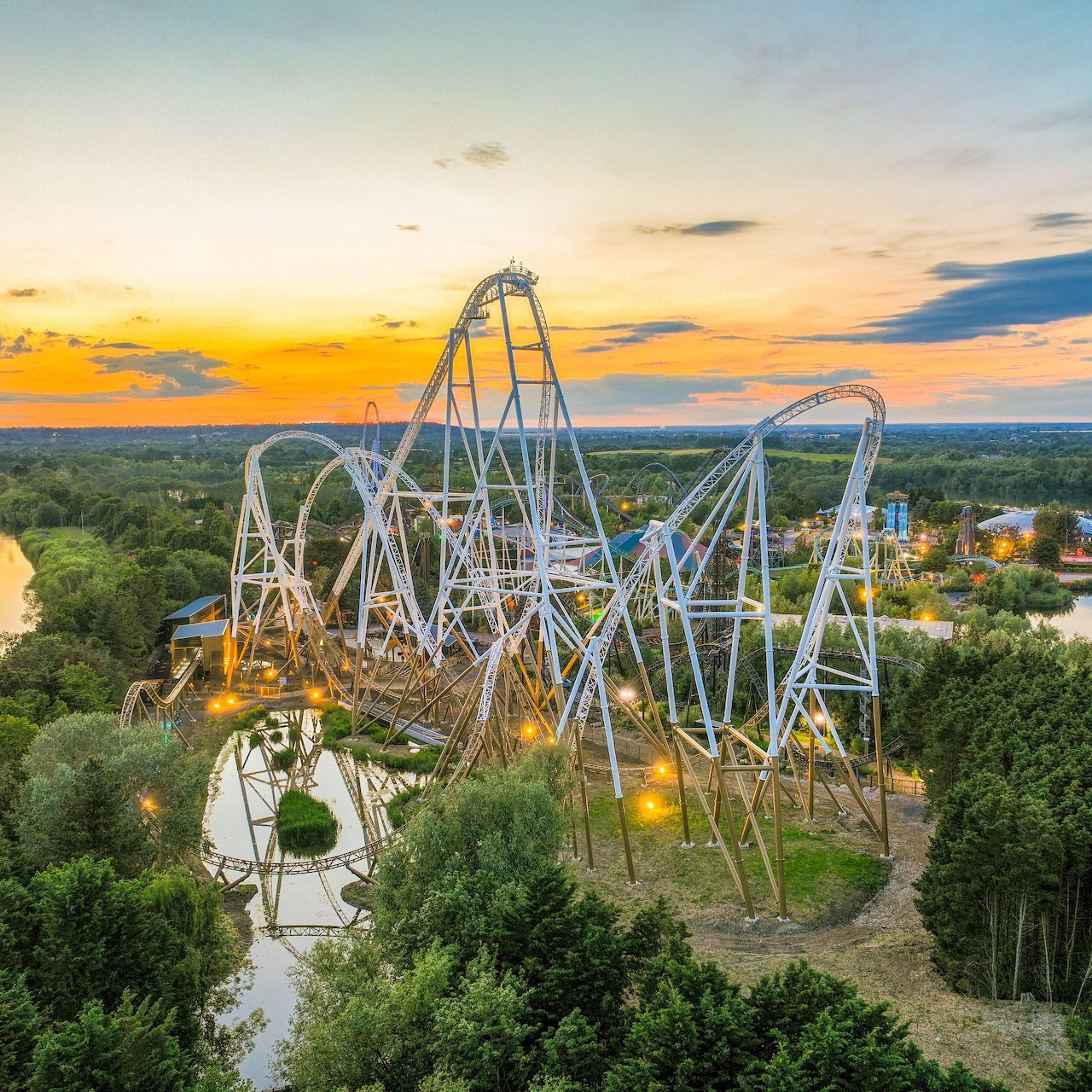 White roller coaster above green trees at sunset with winding tracks and glowing lights
