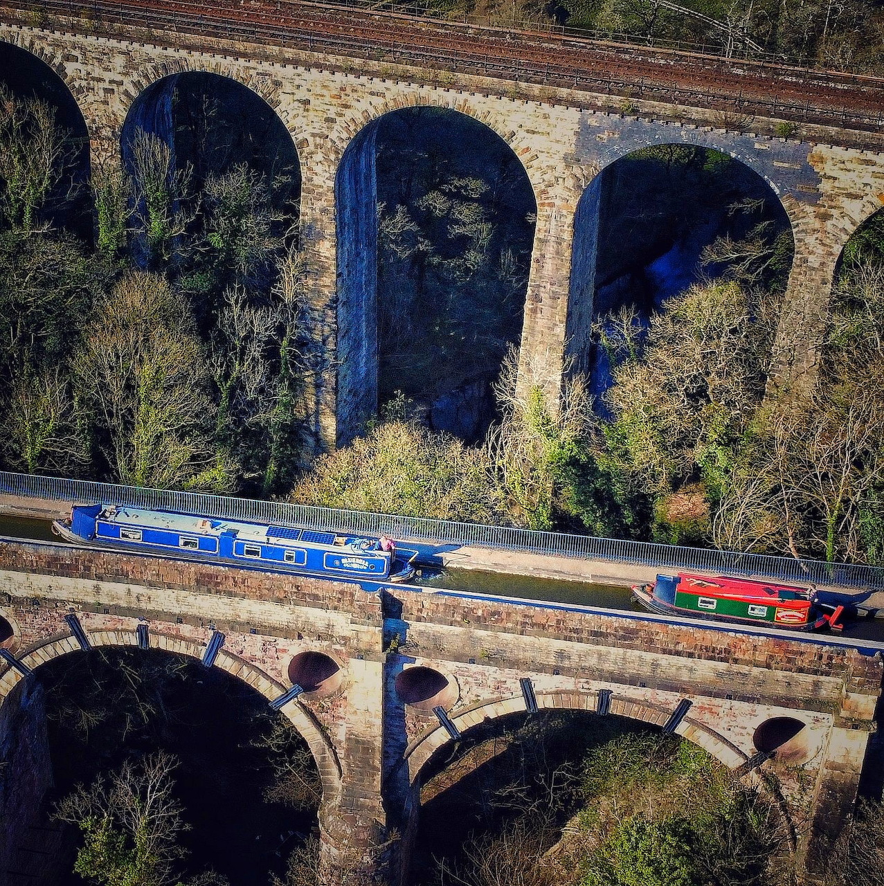 Multi-arch brick aqueduct with blue narrowboat on top canal bridge, surrounded by trees and vegetation