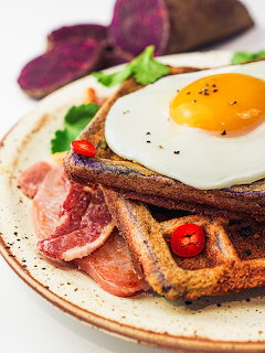 Fried egg on waffle with bacon, chili peppers, purple sweet potato and herbs on a plate