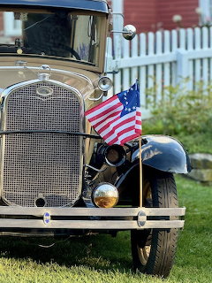 Front of a vintage black and tan car with an American flag on its fender near a white picket fence