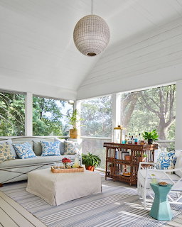Bright sunroom with sectional sofa, coffee table, striped rug, dining table, and large windows overlooking trees