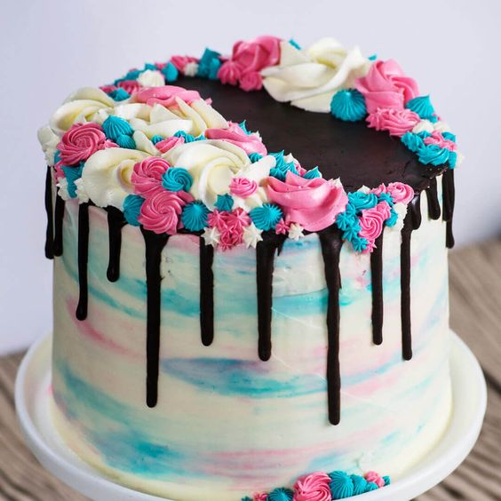 Round birthday cake with pink and blue swirled frosting, chocolate drip, topped with colorful rosettes and meringues