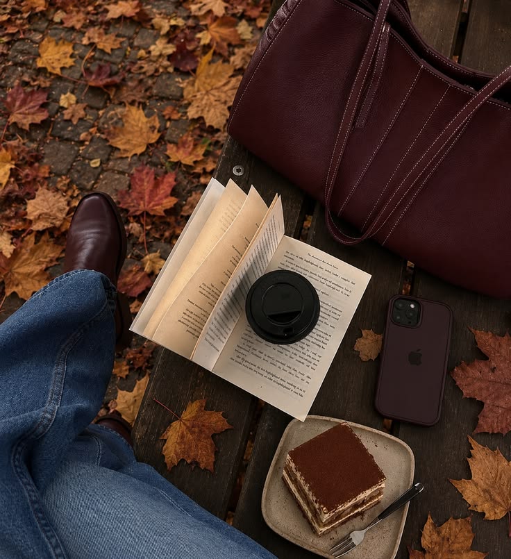 Tiramisu, an open book, coffee, leather bag and phone on a bench amid autumn leaves.