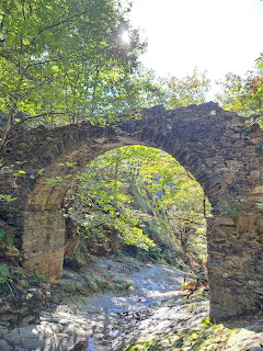 Ancient stone arch bridge over a rocky stream surrounded by green trees in a valley