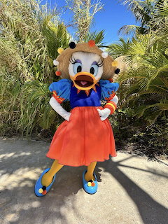 Daisy Duck character in red skirt and blue top posing outdoors with palm trees in background