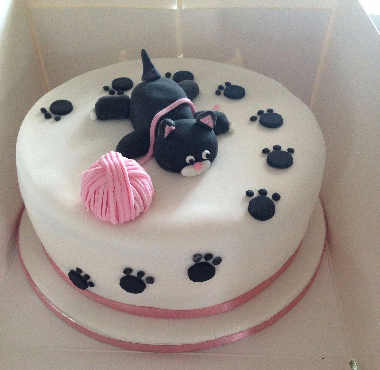White cake decorated with fondant black cat, pink yarn ball, and paw prints