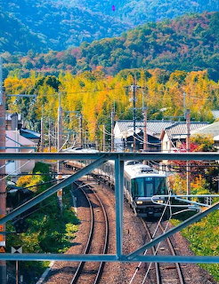 Japanese train at station with autumn-colored trees on mountain hillside seen through metal bridge railing