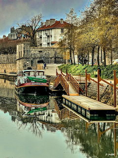Canal boat moored by a dock with old buildings, trees, and reflections in calm water