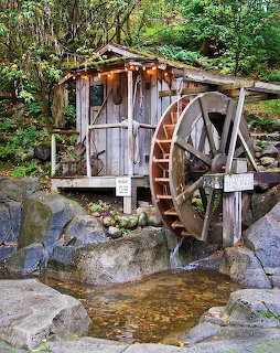 Rustic wooden shed with a large water wheel beside rocks and stream in a lush garden