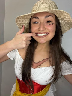 Smiling woman in straw hat pointing to her face with white top
