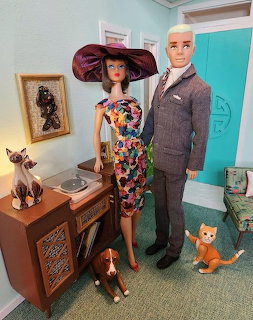 Barbie and Ken dols posed in a miniature room with toy cats, record player, and framed picture