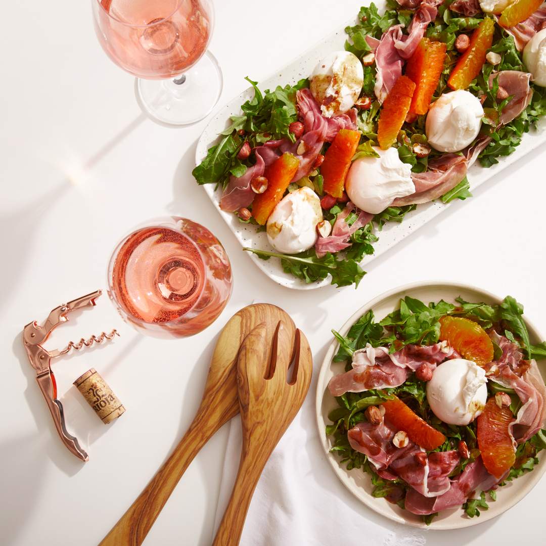 Overhead view of burrata, prosciutto and citrus salad with rosé wine glasses on a white tablecloth