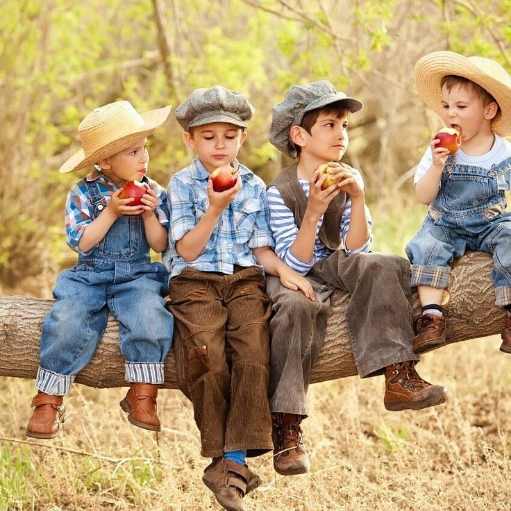 Four children in denim and straw hats sitting on a log eating apples outdoors
