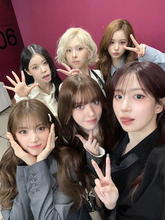 Six young women posing together for a group selfie against a pink wall, making peace signs and hand gestures