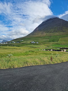 Green mountain towering over a small Faroe Islands village with white houses and lush grass