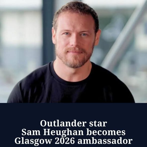 Portrait of Sam Heughan in a black t-shirt smiling at the camera indoors