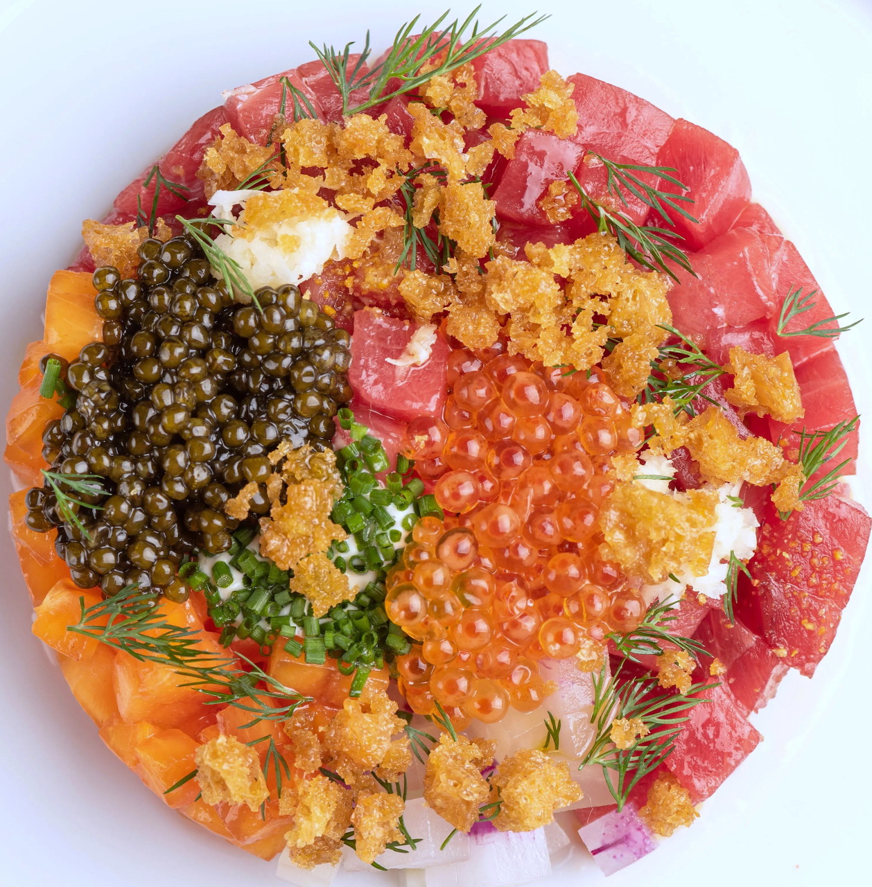 Overhead view of tuna tartare with caviar, salmon roe, herbs, and crispy garnish on a white plate