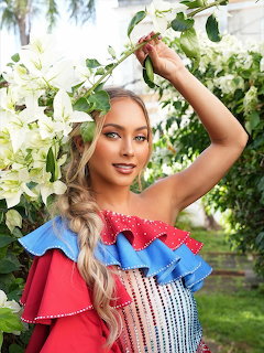 Woman in red, blue, and white ruffled dress reaching up to white flowers in a garden