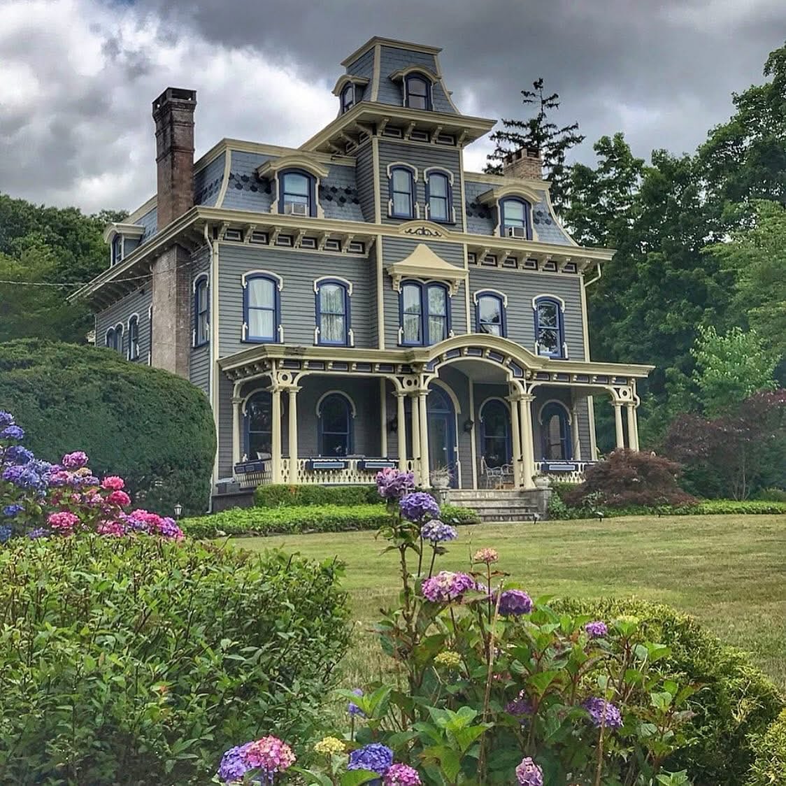 Gray Victorian mansion with ornate details, wraparound porch, and purple hydrangeas in foreground under stormy clouds